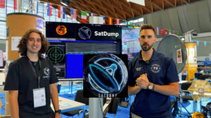 Two presenters at the SatDump booth with spectrum/tracking displays and an L-band dish in the background.