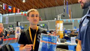 Presenter in a German hamfest hall holding a SatDump information leaflet during a receive-only satellite-decoding demo.